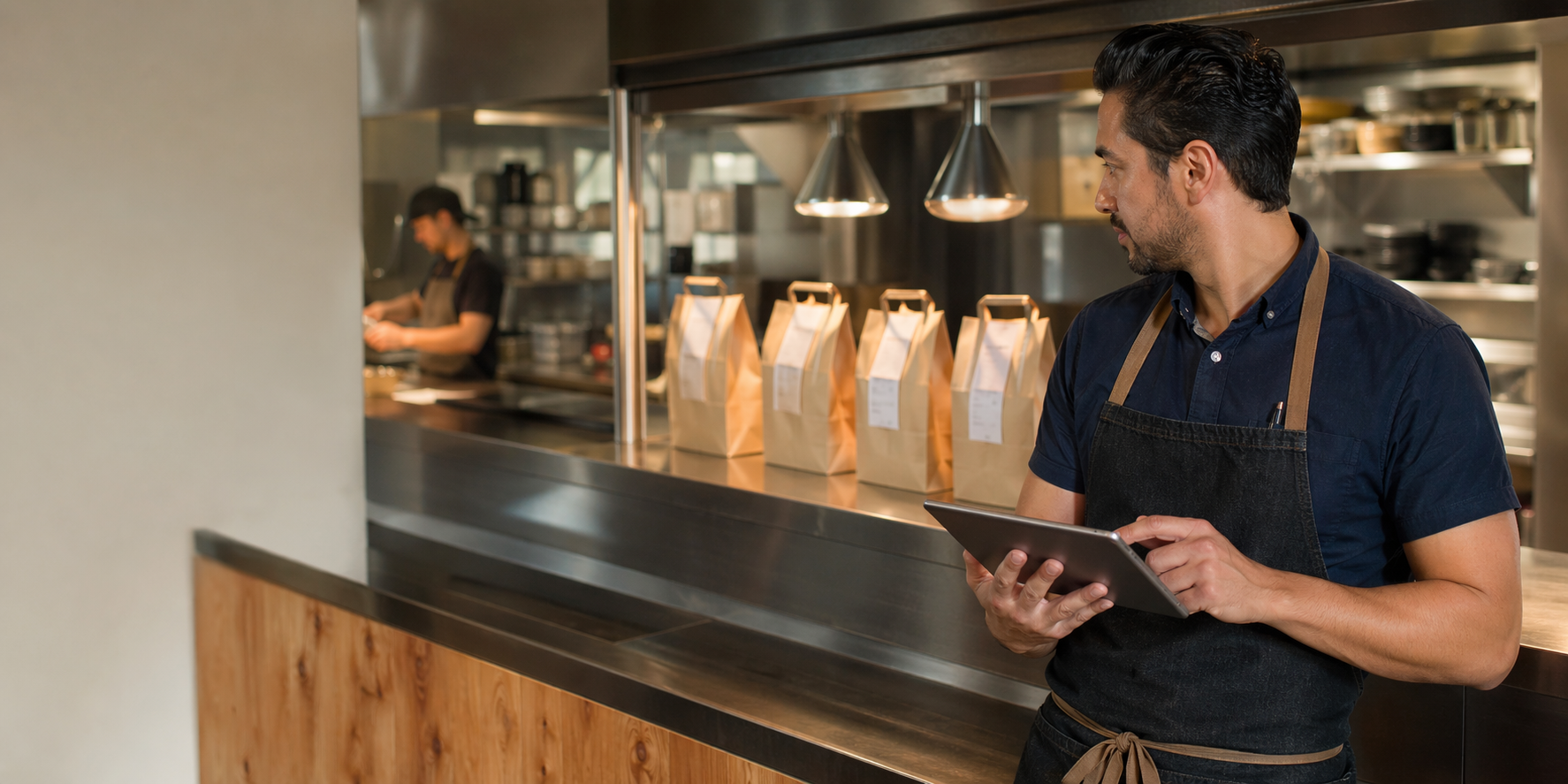 Warm editorial restaurant scene showing a restaurant manager checking a tablet beside ready takeout bags at the kitchen pass.