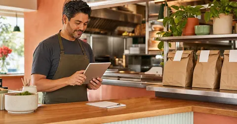 Warm editorial restaurant counter scene showing an owner reviewing online pickup orders on a tablet beside neatly arranged takeout bags on a pickup shelf.